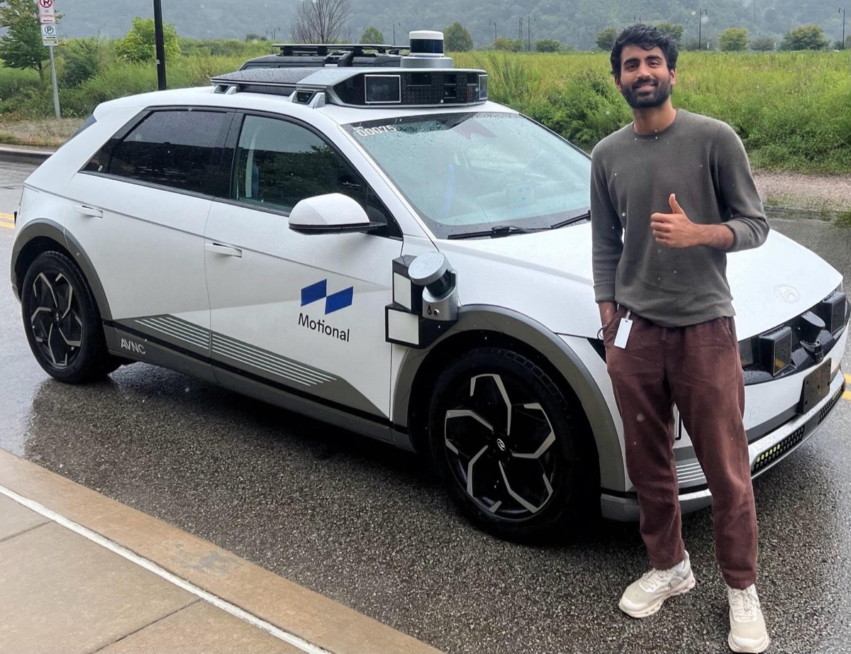 Varun Gandhi giving a thumbs up next to a white Motional IONIQ 5 robotaxi, equipped with lidar and sensor pods, parked on a rainy road in Pittsburgh