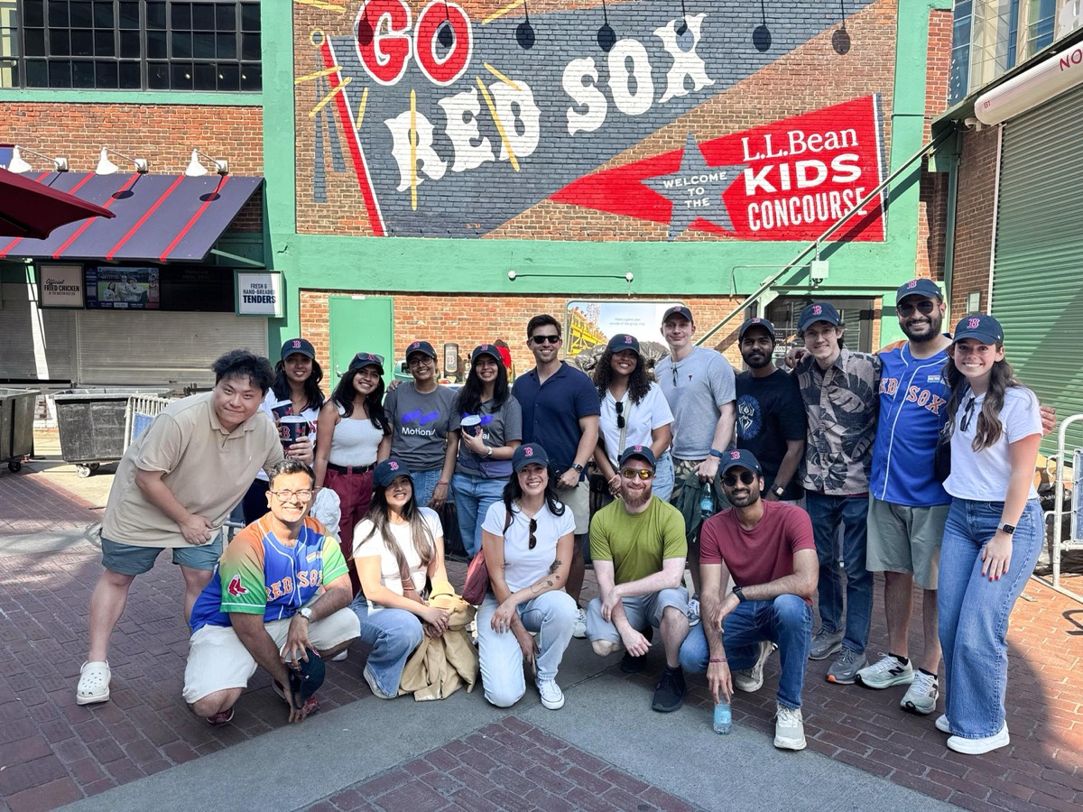 A large group of interns, managers, and team members posing together outside Fenway Park in Boston, in front of a 'Go Red Sox' mural